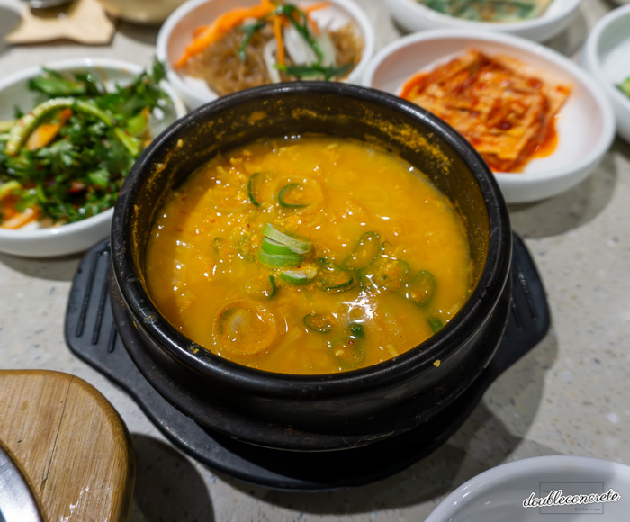 뜨끈한 뚝배기에 담긴 고향 전주비빔밥의 콩비지찌개. 진한 황금빛 국물 위로 다진 파가 올려져 있으며, 뚝배기 주위로 잡채, 겉절이, 나물 등 다양한 반찬이 놓여 있다.