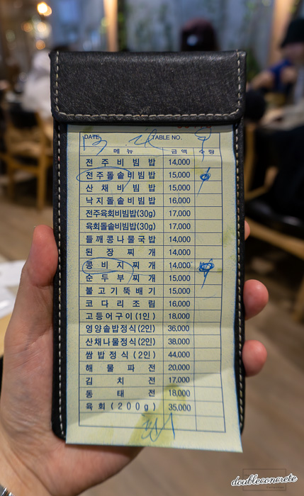 손으로 들고 있는 검은 가죽 커버의 계산서 속, 한식당 '고향 전주비빔밥'의 메뉴 및 가격이 적혀 있다. 주문된 항목에는 체크 표시가 되어 있다.