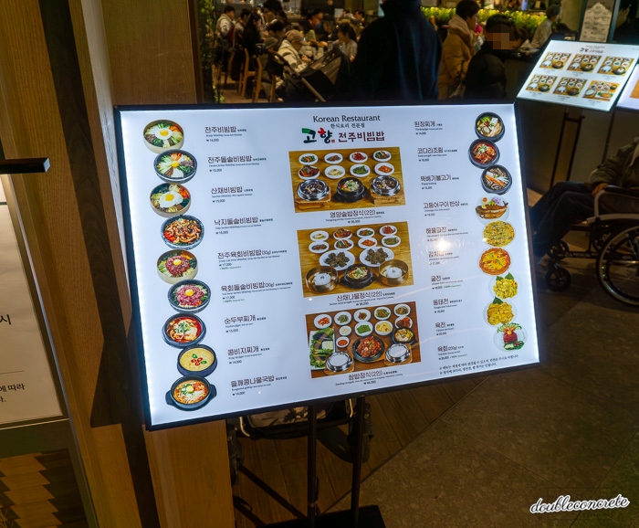 현대백화점 킨텍스점 내 한식당 '고향 전주비빔밥'의 메뉴판. 비빔밥, 찌개류, 정식 메뉴 등 다양한 한식 요리가 표시된 메뉴 보드가 조명이 들어간 형태로 전시되어 있다. 메뉴판 뒤로는 식당 내부와 손님들로 붐비는 모습이 보인다.