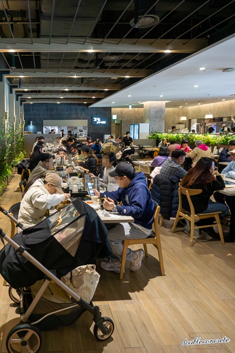 현대백화점 킨텍스점 8층에 위치한 한식당 ‘고향 전주비빔밥’의 내부 모습. 점심시간대 많은 손님들이 가득 차 있으며, 유모차를 둔 가족, 연인, 친구들이 한식을 즐기고 있다. 매장은 넓고 개방적이며, 벽면의 초록 식물 장식과 조명이 따뜻한 분위기를 연출한다.