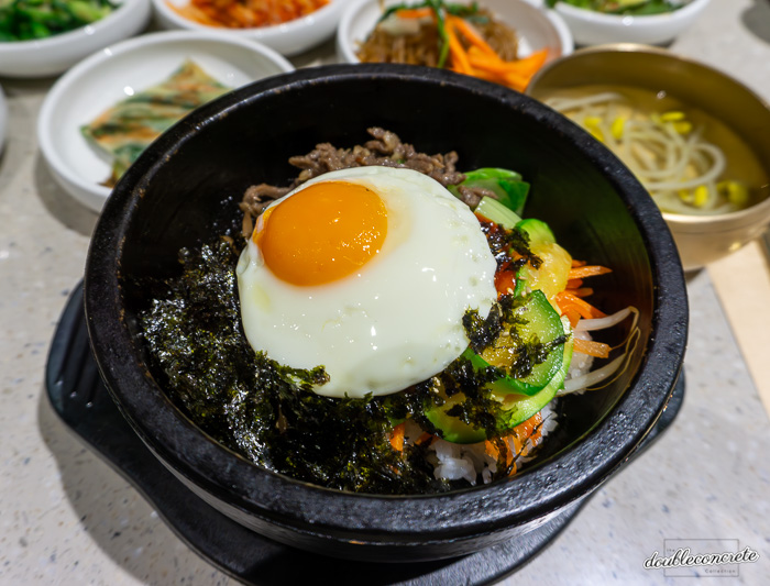 돌솥에 담긴 전주비빔밥 한 그릇. 윤기 나는 계란 프라이가 올려져 있으며, 다양한 채소와 고기, 김 가루가 조화를 이루고 있다. 주변에는 다양한 반찬과 콩나물국이 함께 제공된다.