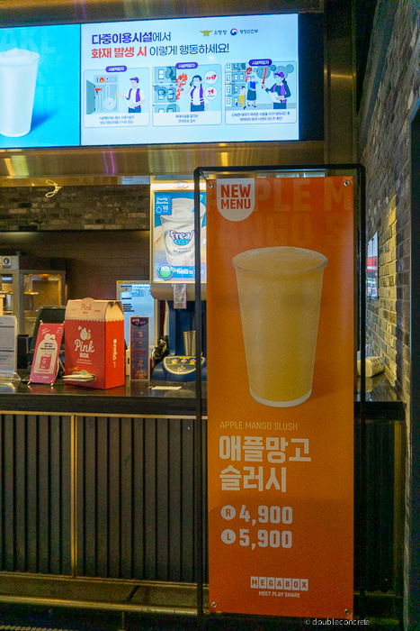 메가박스 매점에서 새롭게 출시된 애플망고 슬러시 홍보 배너가 세워져 있다. 배너는 오렌지색 배경에 투명한 컵에 담긴 노란빛의 슬러시 이미지가 강조되어 있으며, 가격은 R 사이즈 4,900원, L 사이즈 5,900원으로 표시됨.배경에는 팝콘 박스, 아이스크림 기계, 그리고 상단에는 다중이용시설에서 화재 발생 시 대피 요령을 안내하는 디지털 스크린이 설치되어 있다.
