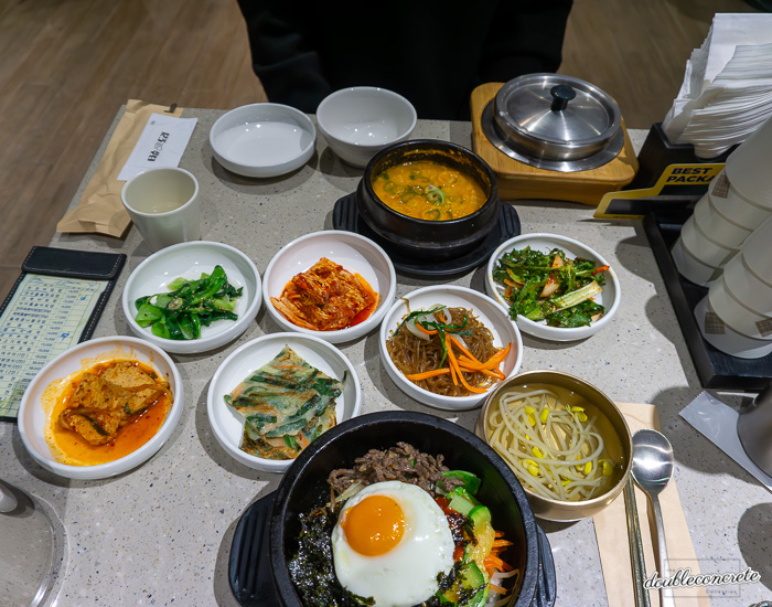 뜨거운 돌솥에 담긴 전주비빔밥과 콩비지찌개, 그리고 다양한 반찬이 함께 제공된 한식 한상차림. 반찬으로는 김치, 잡채, 김치전, 오이무침, 콩나물국 등이 제공되며, 따뜻한 밥이 뚝배기에 담겨 나온다.