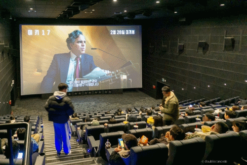 메가박스 돌비 애트모스 상영관에서 미키17 (Mickey 17) 예고편이 상영되는 모습. 큰 스크린에 로버트 패틴슨이 등장하는 장면이 나오고 있으며, 관객들이 좌석에 앉아 영화 시작을 기다리고 있다. 어두운 극장 내부와 함께 팝콘과 음료를 들고 있는 관객들의 모습도 보인다.
