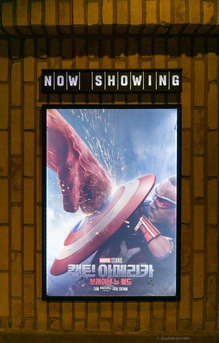 벽면에 부착된 "NOW SHOWING" 조명 아래, 마블 스튜디오의 '캡틴 아메리카: 브레이브 뉴 월드' 공식 포스터가 걸려 있다.포스터 속 장면은 **새로운 캡틴 아메리카(샘 윌슨)**가 레드 헐크의 강렬한 주먹을 방패로 막아내는 순간을 담고 있으며, 충돌로 인해 방패에서 불꽃이 튀는 역동적인 연출이 돋보인다. 하단에는 개봉일 정보와 함께 2월, 최초 개봉, 극장 대개봉이라는 문구가 삽입되어 있음.