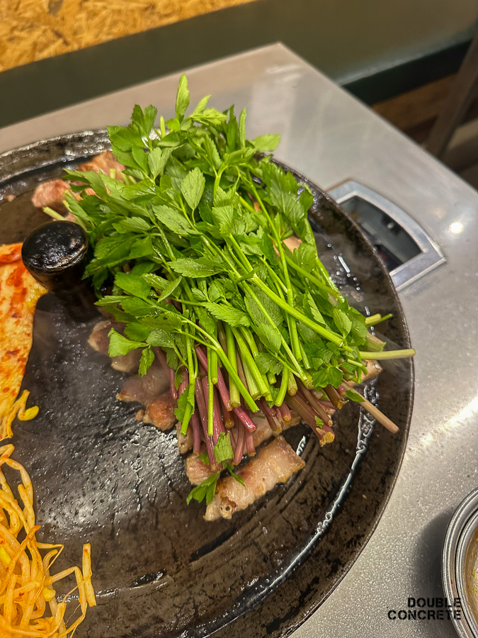 삼겹살 위에 신선한 미나리가 가득 올려진 모습. 미나리가 고기와 함께 구워지는 장면.