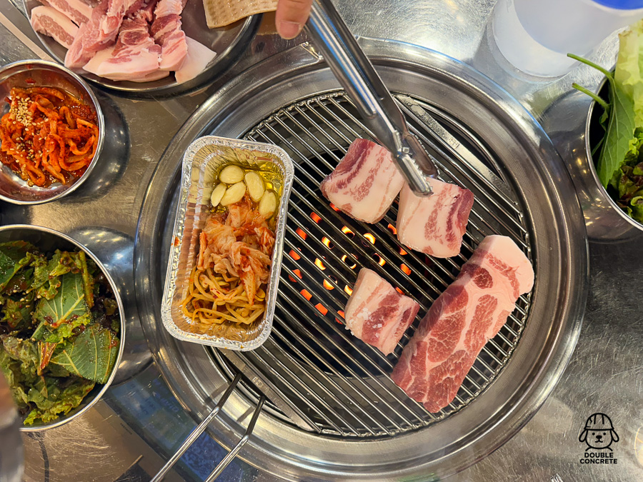 연탄불 위에 삼겹살과 항정살, 알루미늄 접시에 반찬 구성이 올라간 모습